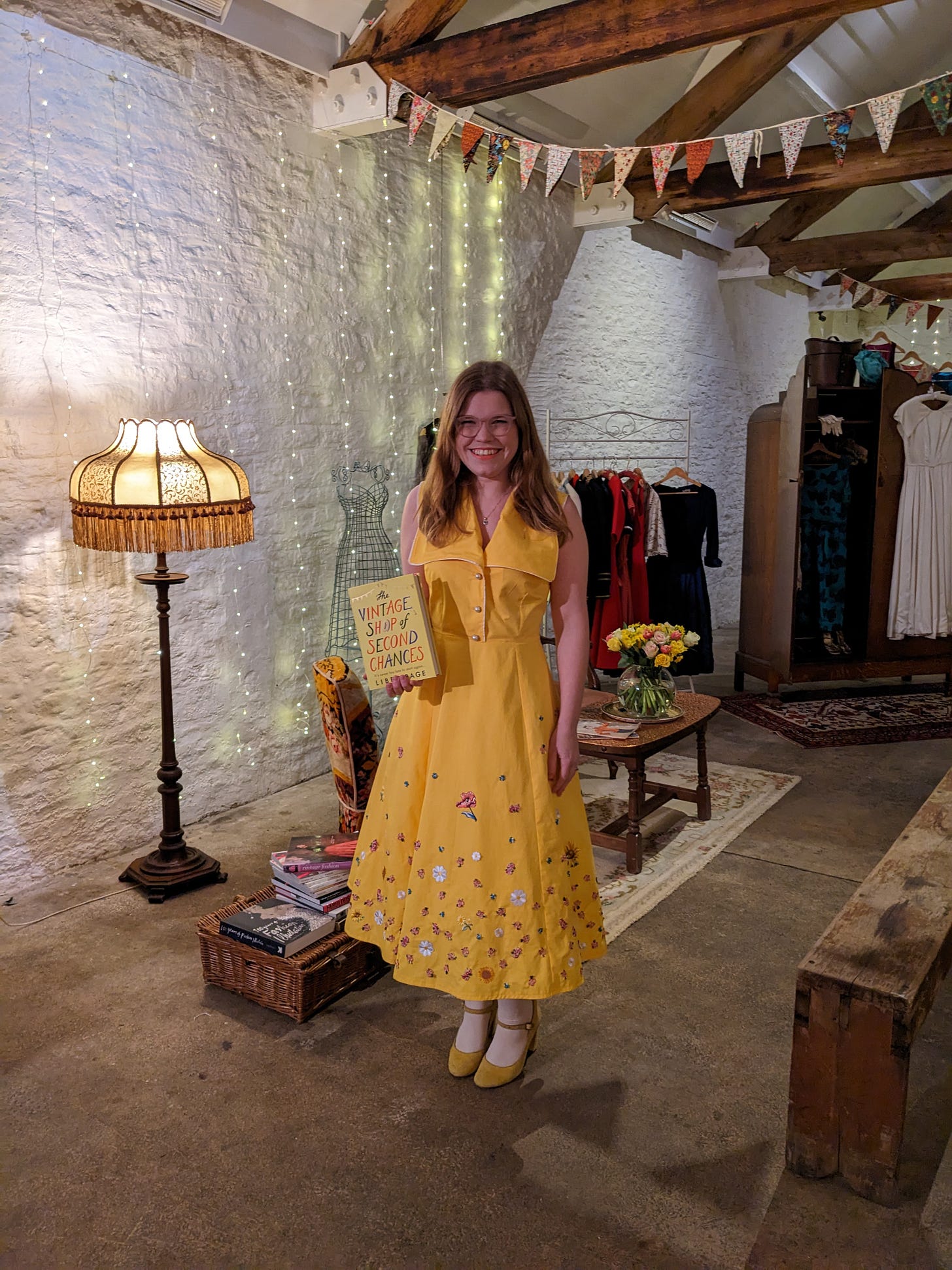 Libby wearing a yellow vintage style dress covered in flowers, holding a copy of her book The Vintage Shop of Second Chances.