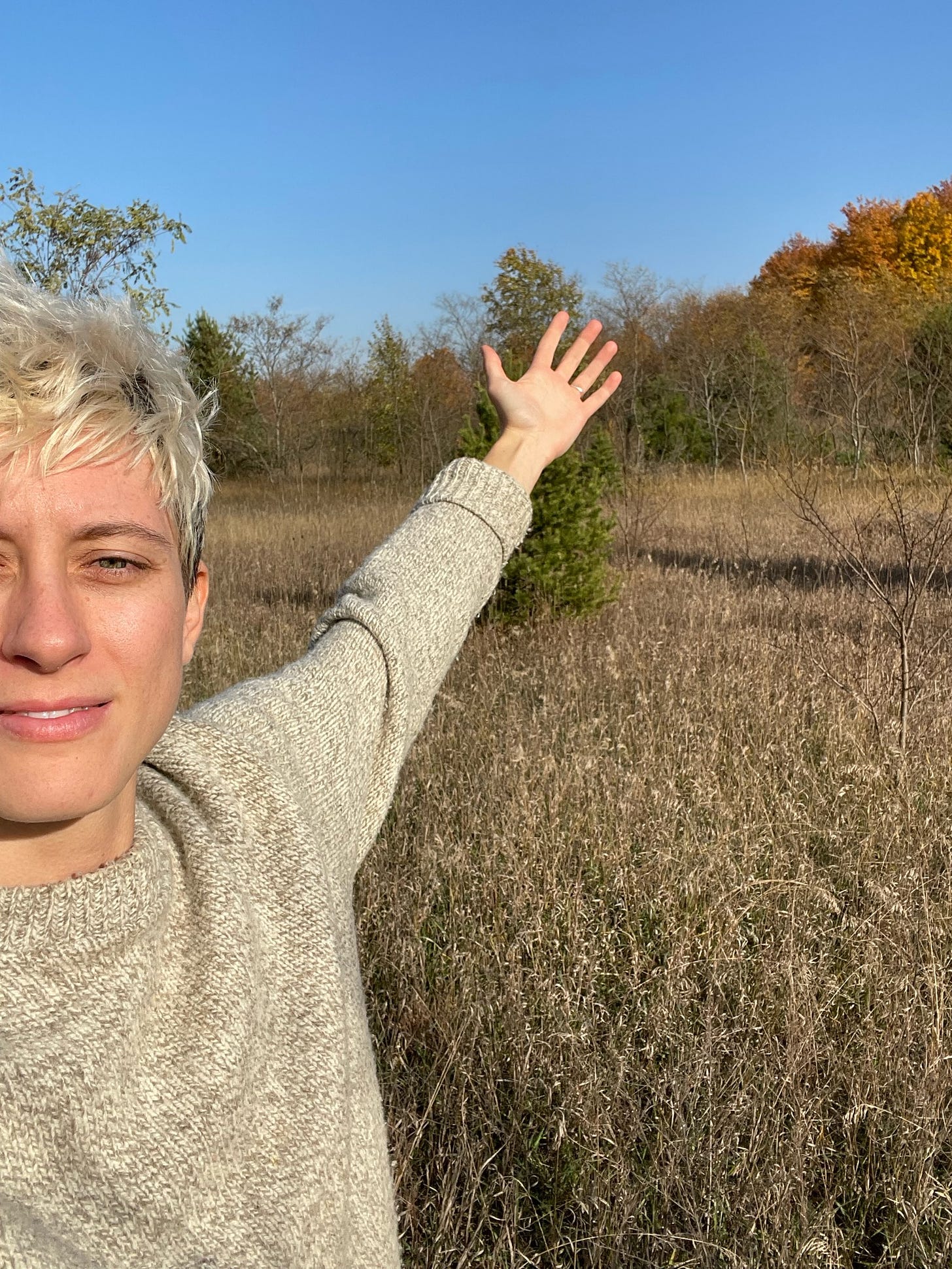 Elle smiling at camera with hand extended in front of fall foliage property.