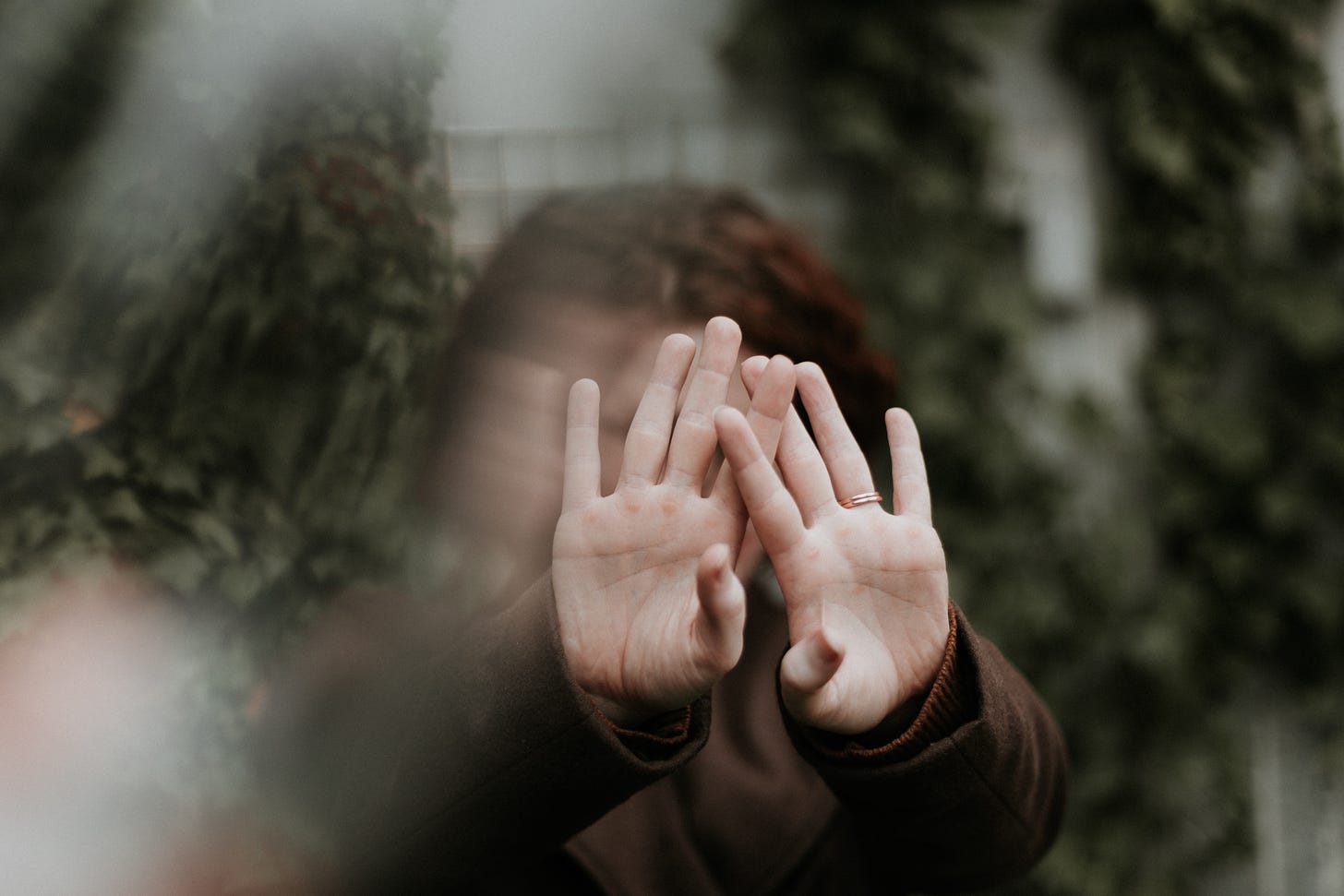 Two hands are palms out towards the camera, with a few fingers intertwined. The background is blurry with some greenery and the face of the person whose hands are outstrecthed.