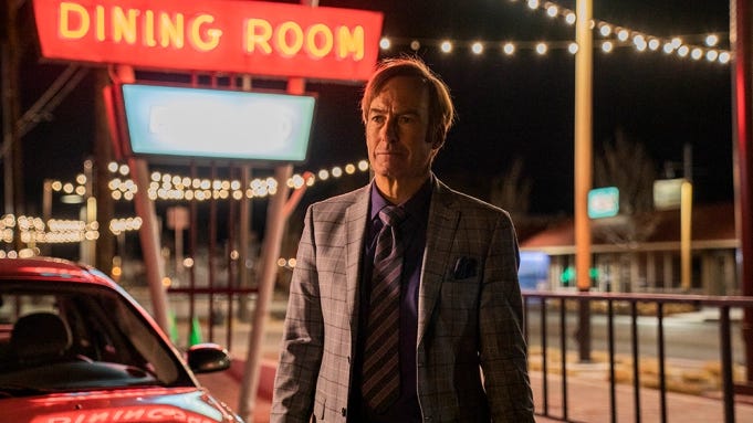 Bob Odenkirk as Jimmy McGill in a suit standing in front of a neon sign that reads Dining Room, in the series Better Call Saul.