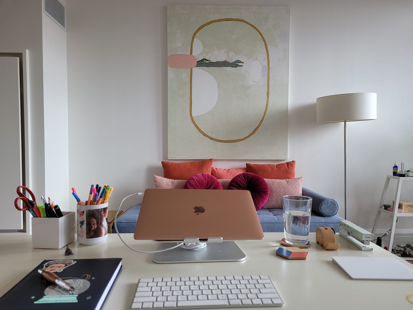 A desk with a laptop, notebook, two pencil ups, a glass of water, and a small crystal in the foreground. A couch with pillow, a large abstract painting, a floor lamp, and a bookshelf with crystals in the background.