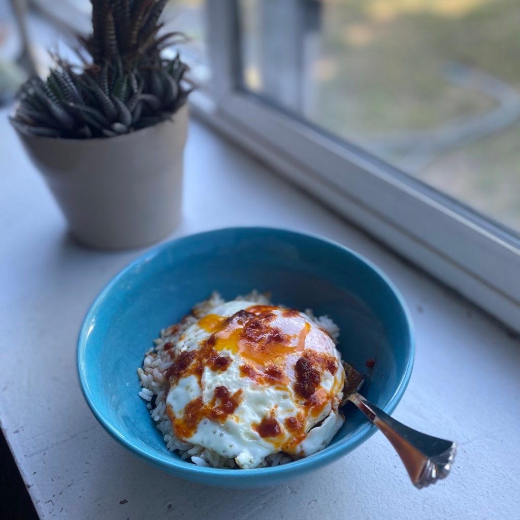 A blue bowl of fried rice topped with an over easy egg that's been split open at the top. Yolk drips down from the crack in the white, and chili miso dots the top. The bowl is on a windowsill with a plant in the background.
