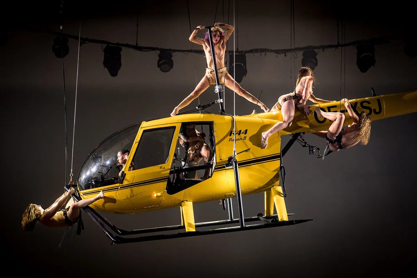 Imagem: Helicóptero amarelo pendurado por hastes. Uma mulher nua no alto em pé, duas dentro, uma pendurada no bico frontal e duas penduradas na parte detrás do helicóptero.