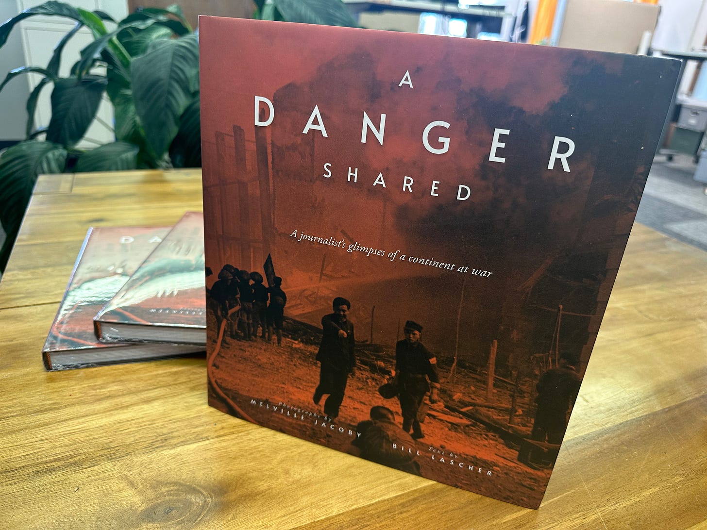 A copy of the book A Danger Shared with photographs by Melville Jacoby and text by Bill Lascher stands on a wooden table with two more shrink-wrapped copies of the book resting on the table behind it.