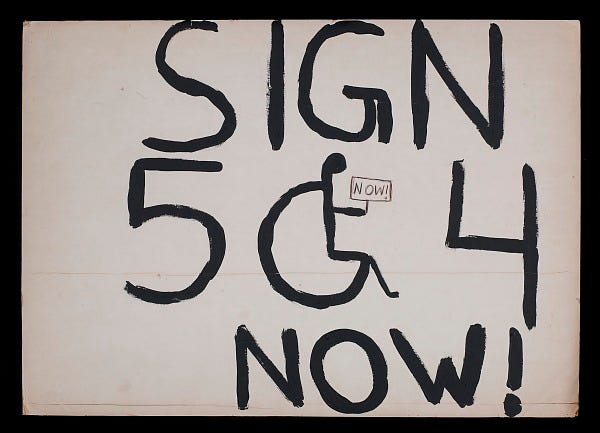 Hand-written letters on white background saying "Sign 504 now!" The zero in the number "504" is the handicapped symbol (person in wheel chair).