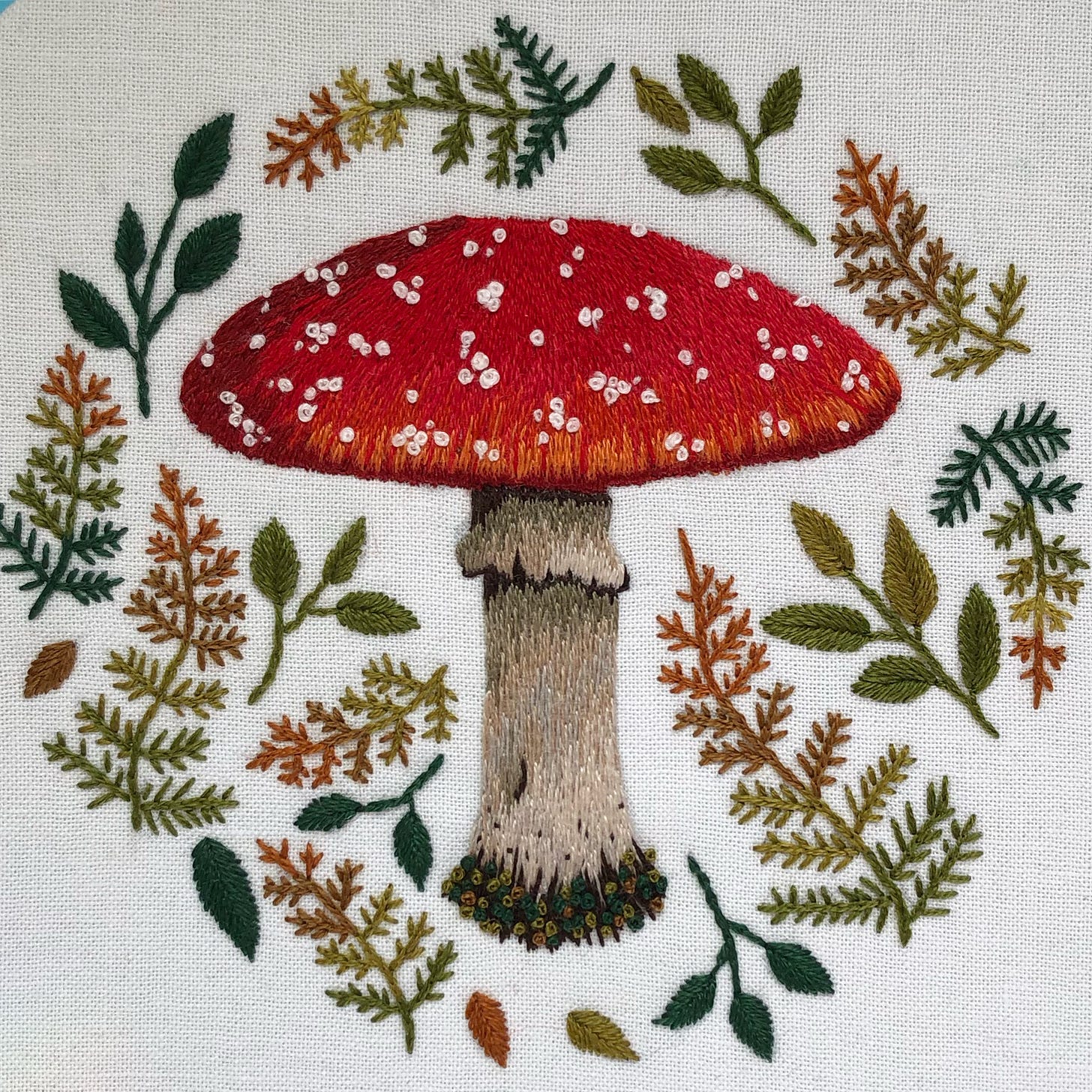 An embroidery of a red-capped mushroom surrounded by leaves and ferns