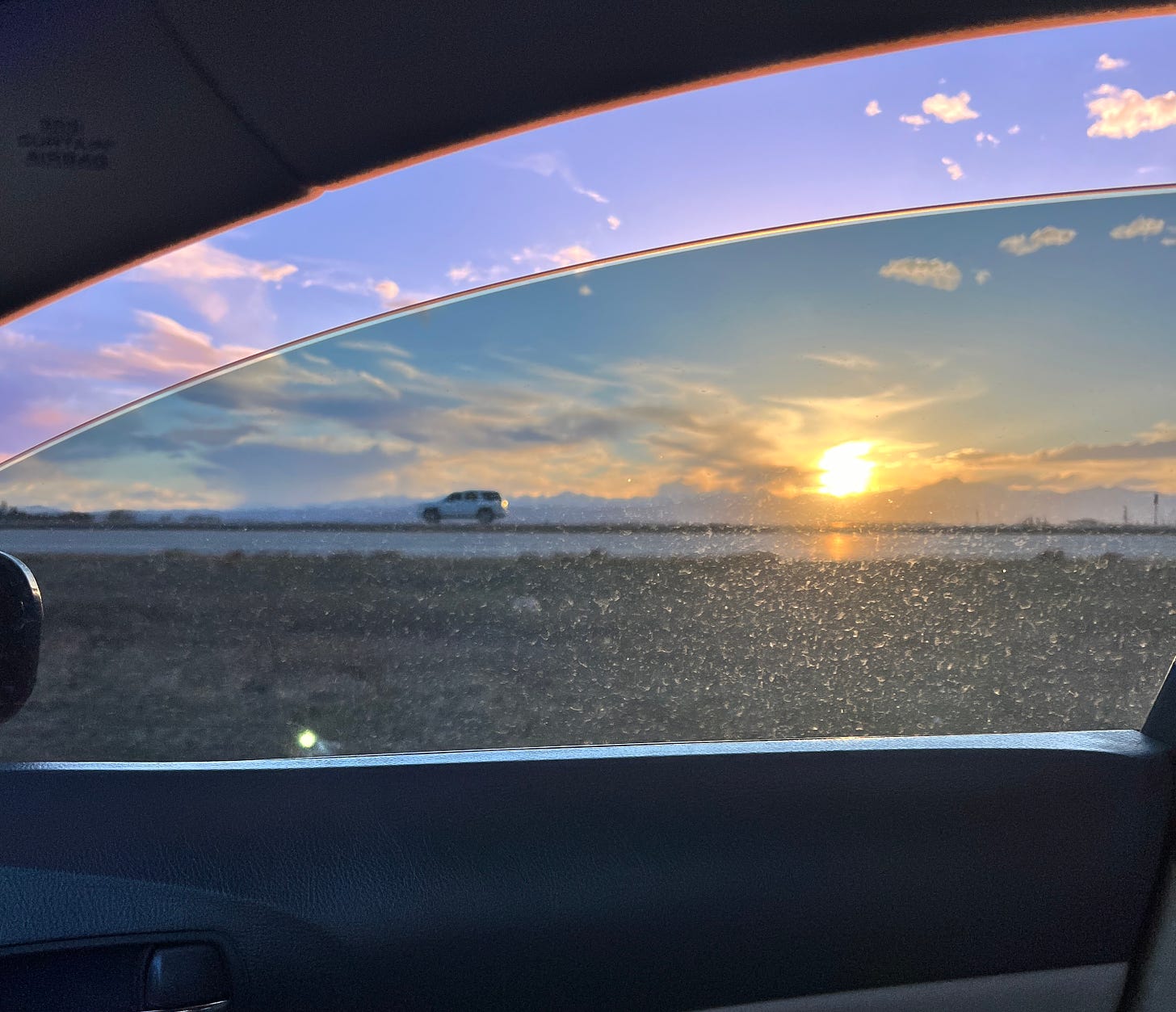 photo looking out of a car window shows the sun settings over the mountains with clouds in the sky and a white SUV driving in the same direction