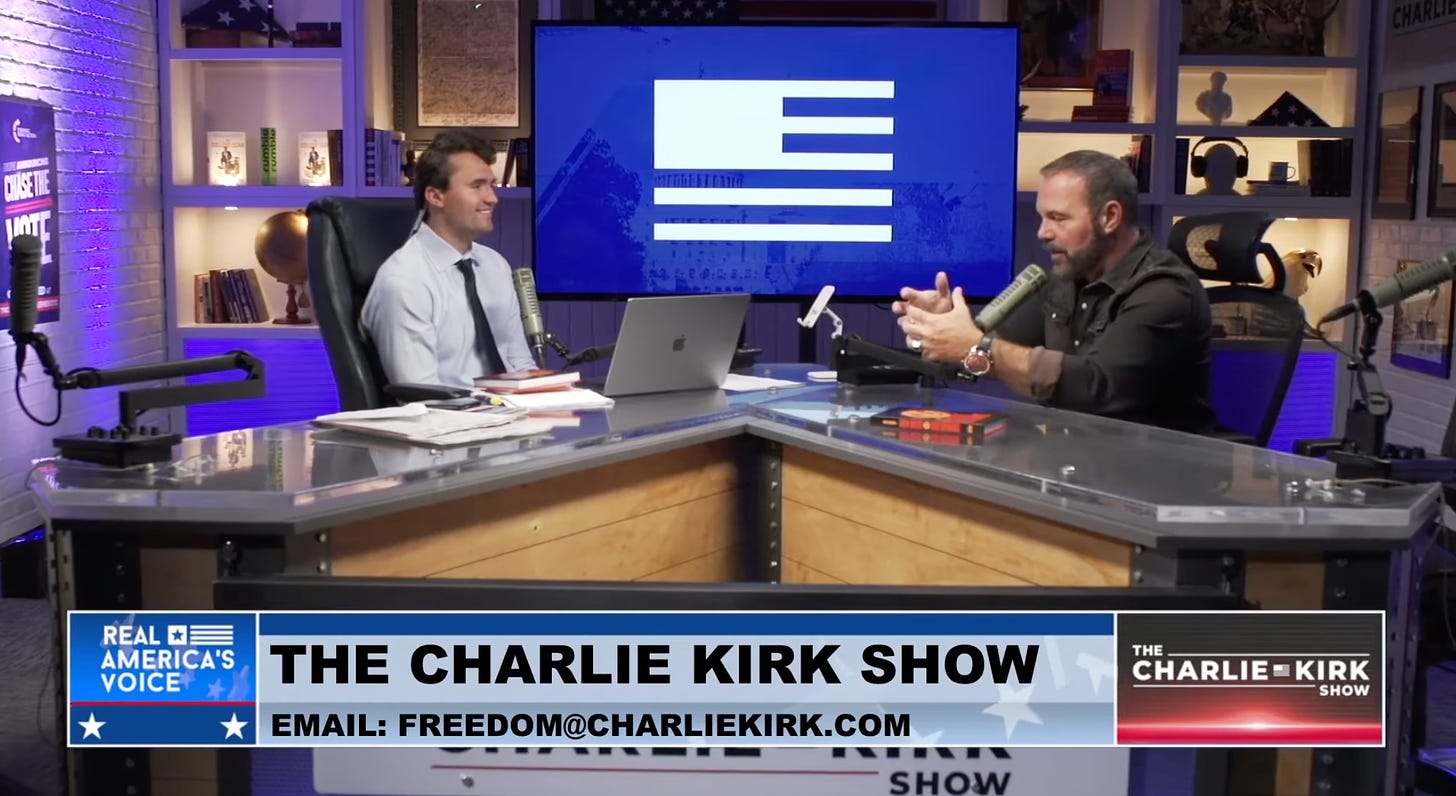 Charlie Kirk and Pastor Mark Driscoll sitting across from one another on the set for the Charlie Kirk show. Driscoll is speaking into the mic and gesticulating with his hands. One of his many crappy books sits on the table. 