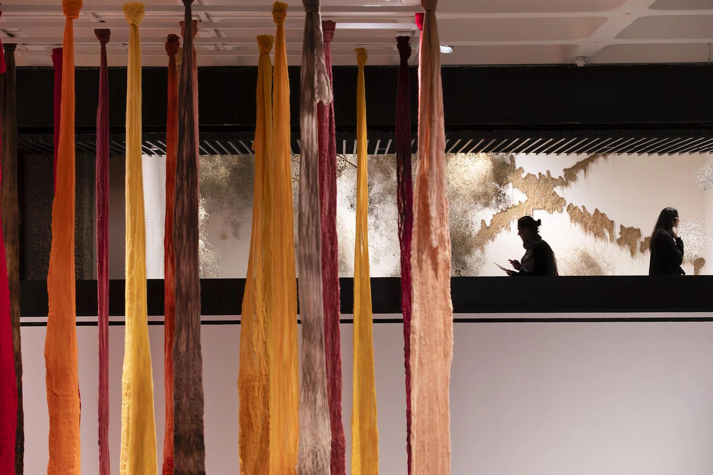 View of the upstairs of Barbican’s 2024 Unravel exhibition, showing Cecilia Vicuña’s hanging coloured felt in the foreground, and Igshaan Adams’ large-scale wall work in the background [Credit: Barbican].