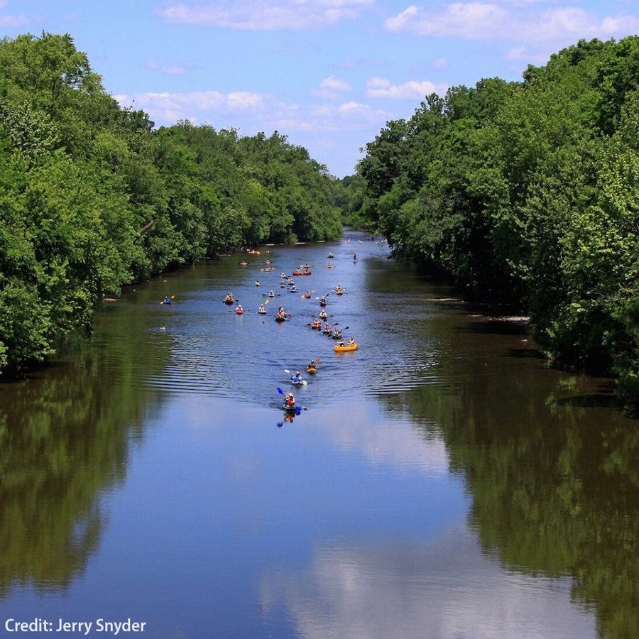 Paddlers in kayaks and canoes paddle down the Perkiomen Creek. Green trees line both banks.