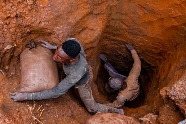 A worker hauls a bag of raw cobalt from a hole in the ground. Another worker stands deeper in the hole.