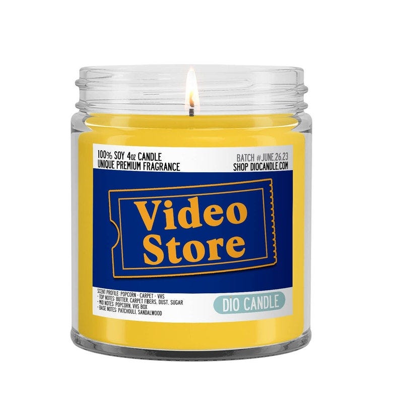 May include: A yellow soy candle in a clear glass jar with a black and blue label that reads 'Video Store'. The label also includes the text '100% Soy 4oz Candle', 'Unique Premium Fragrance', 'Batch #June 26.23', 'Shop Diocandle.com', and 'Dio Candle'. The scent profile is described as 'Popcorn-Carpet-VHS' with notes of butter, carpet fibers, dust, sugar, popcorn, VHS box, patchouli, and sandalwood.