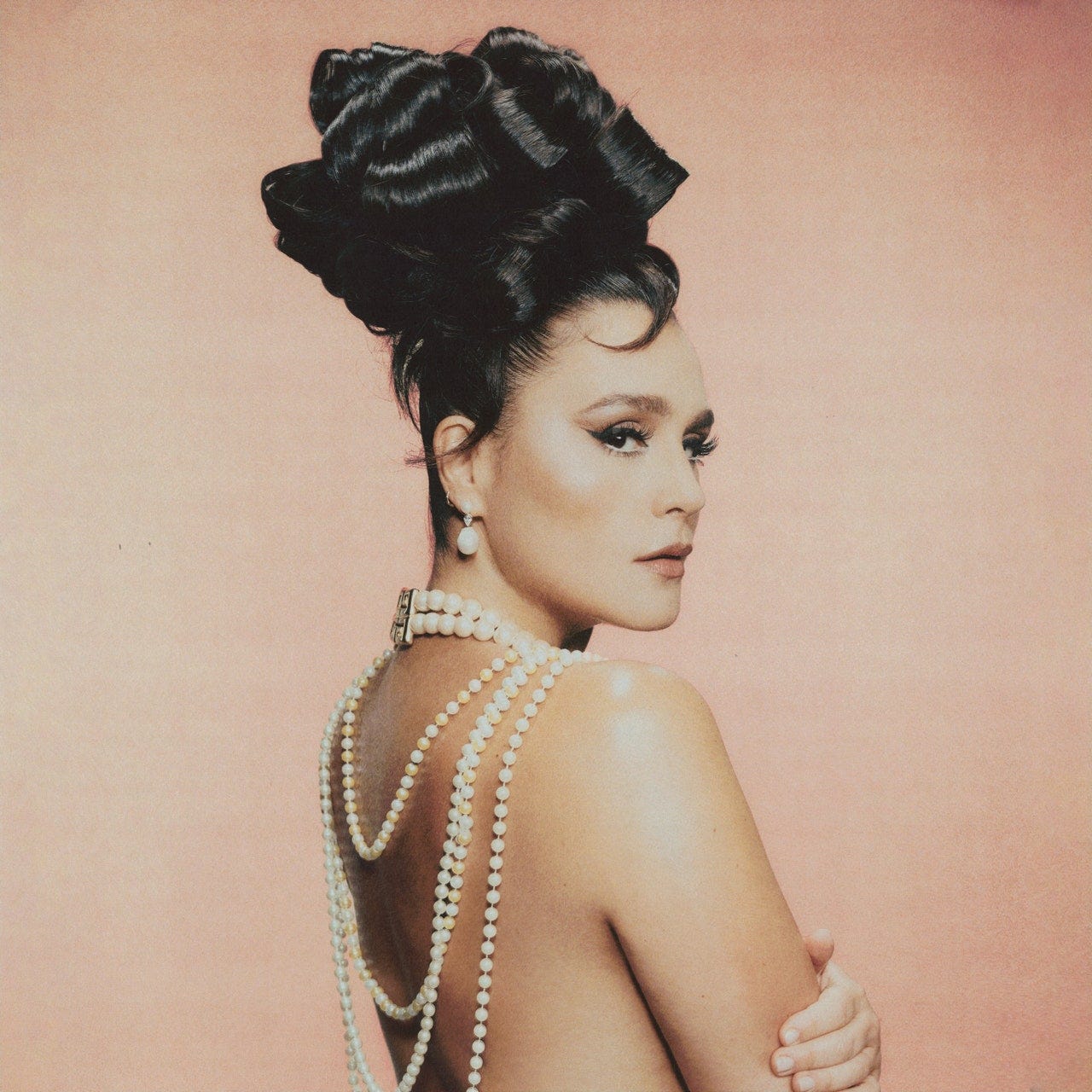 Jessie Ware stands with her back slightly to the camera wearing just a few strands of pearls that drape over her back and an elaborate updo.