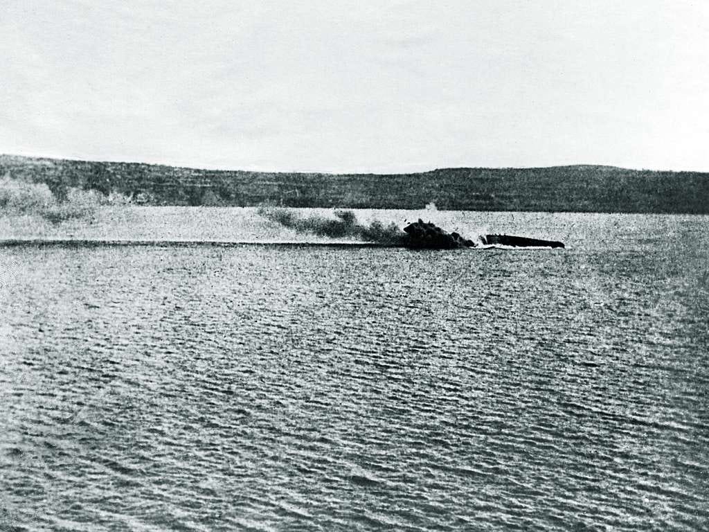 Grainy black and white photo of a ship's keel lying low in the water. Smoke drifts away from the hull. Low hills are in the bachground.