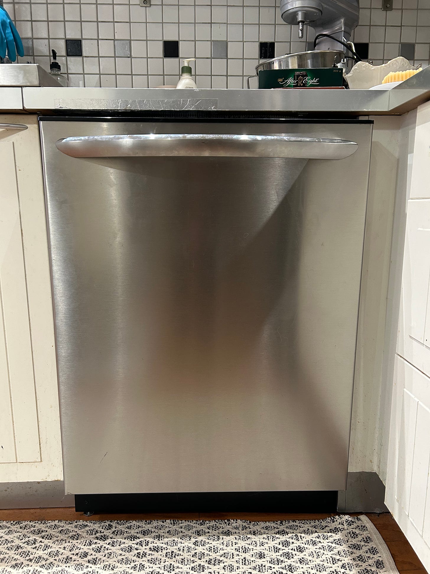 A stainless steel dishwasher sits installed beneath a grey countertop in a kitchen. on the wall behind the dishwasher is a series of white square backsplash tiles with grey and black tiles at set intervals. 