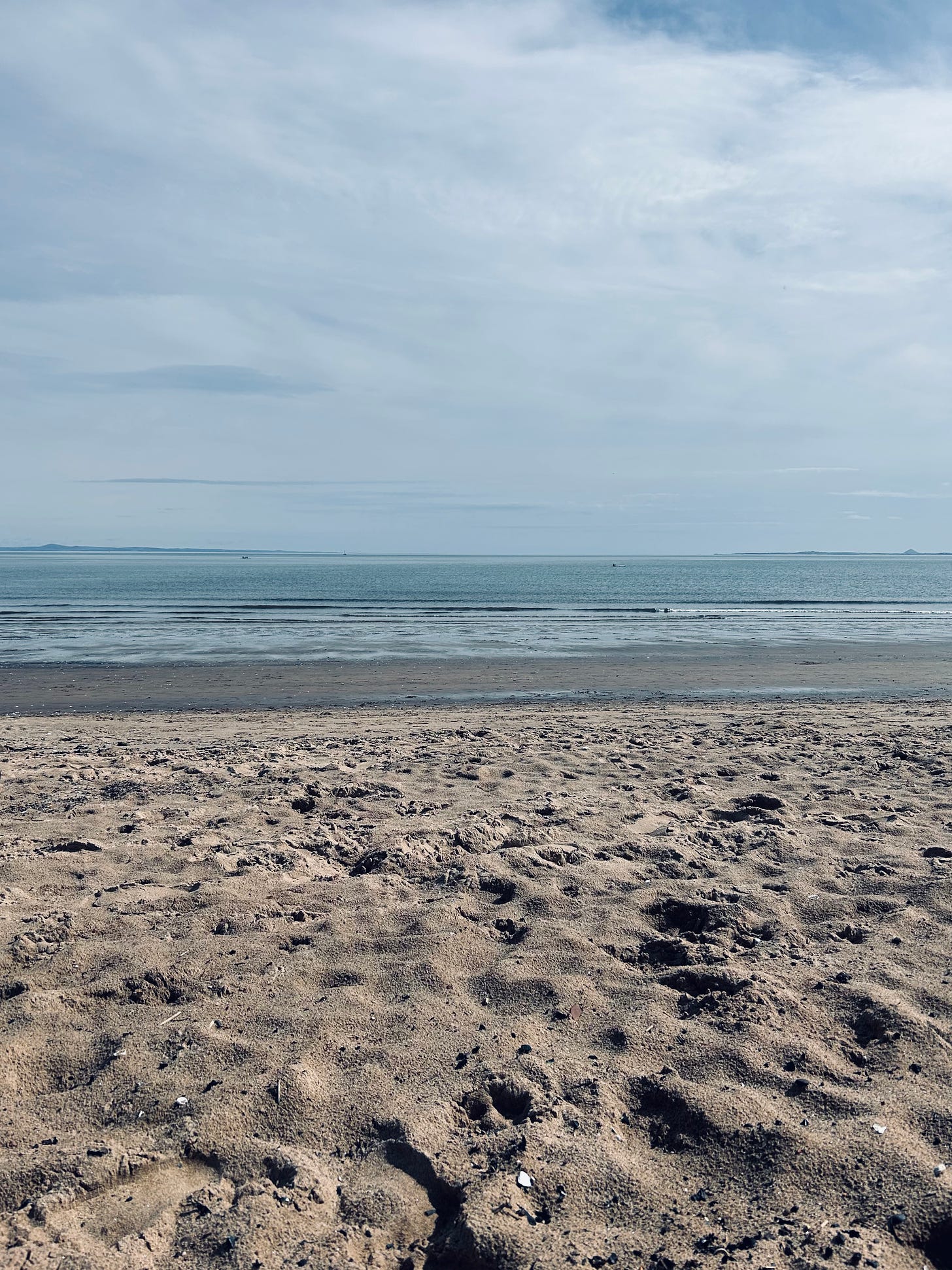 A photo of the Portobello Beach coast. In the foreground is a sandy shore. Then a layer of wet sand. At the horizon is blue sea water, topped off with blue, cloudy skies.