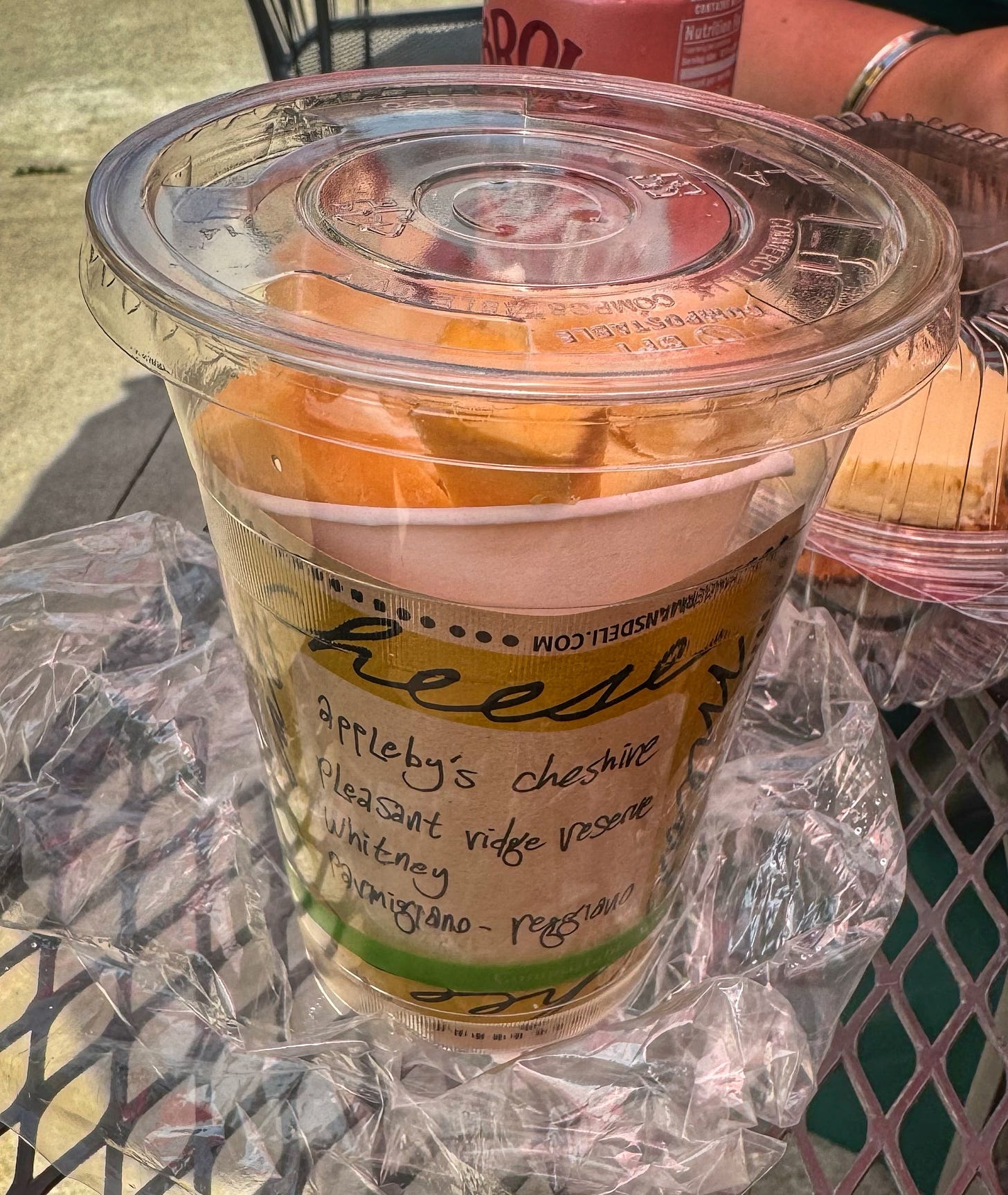 A plastic cup containing a "cone of cheese. as sold by Zingerman's. There are a few chunks of several different kinds of cheese. A label inside the cup lists the varieties as Appleby's Cheshire, Pleasant Ridge reserve, Whitney, and Parmigiano Reggiano.