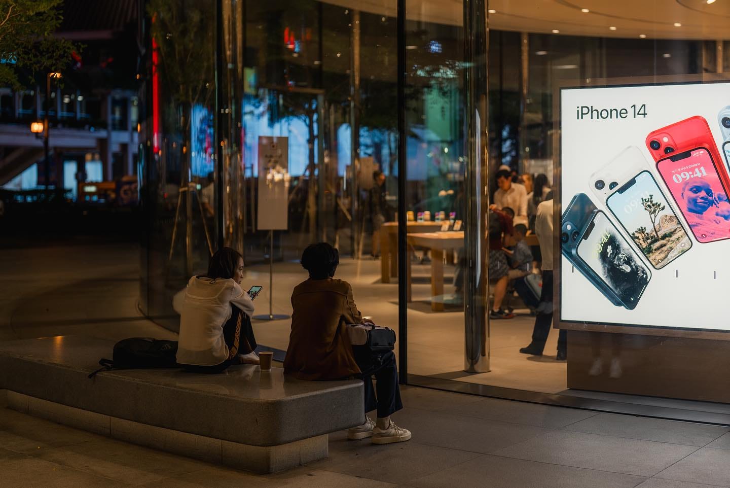 Two people sit on a stone bench outside Apple Central World at night.