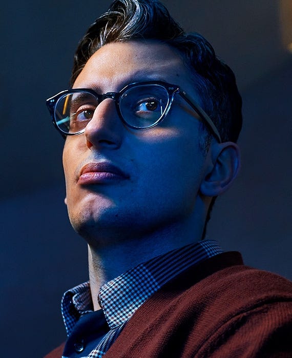 Ryan J Haddad wearing a red sweater and glasses in front of a dark background