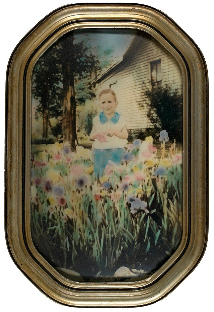 A photographic print with applied color from the 1920's-30's of a young white boy standing among flowers, with a house in the background.