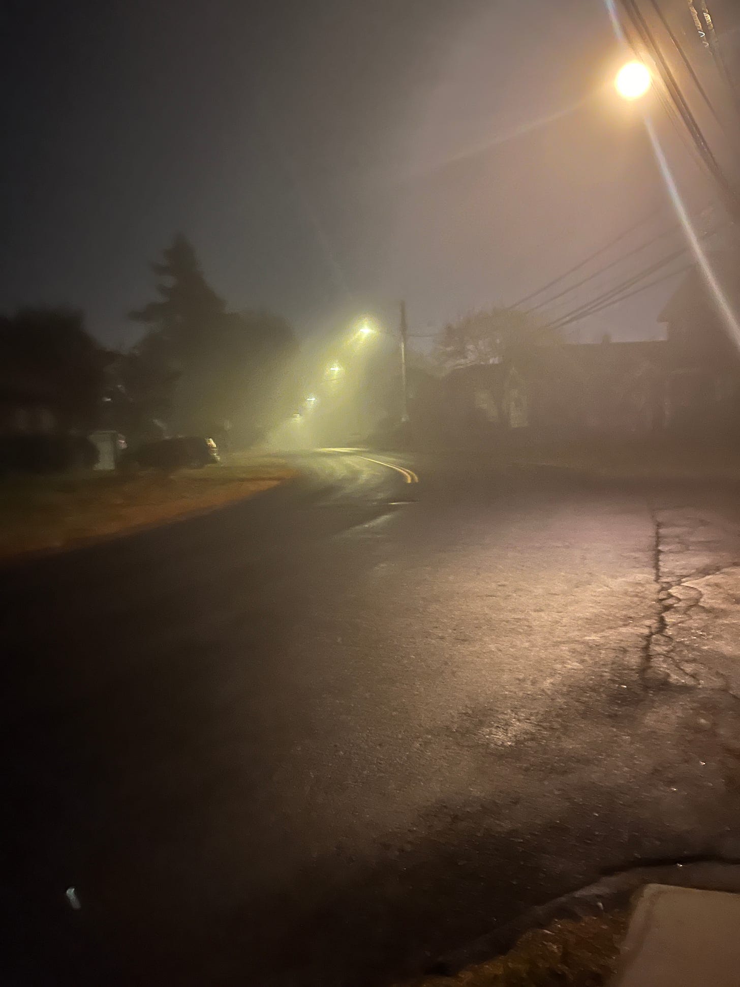 A roadway curving to the left, with powerlines above, in the darkness, and the streetlights in fog trace a trail.