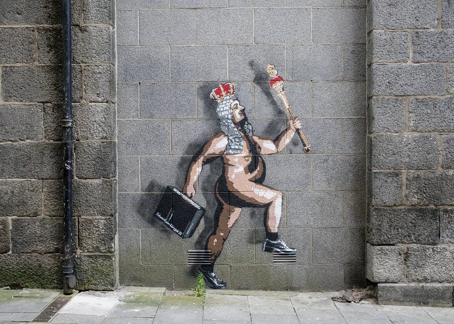 This image captures a piece of street art by Edward von Lõngus titled 'The Naked Emperor'. The artwork, spray-painted on a grey urban wall, depicts a figure resembling a traditional emperor, but humorously presented without clothes. The character has a long beard, a red crown, and holds a sceptre aloft. Instead of regal attire, the emperor sports black shoes with thick soles. The figure appears to be stepping off a pair of black blocks, giving the impression of motion. A drainpipe bisects the image, adding to the urban feel of the scene.