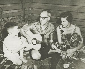 Photo of Hermes Nye sitting on the floor with his wife and young child. Hermes is playing the guitar, and they all look happy