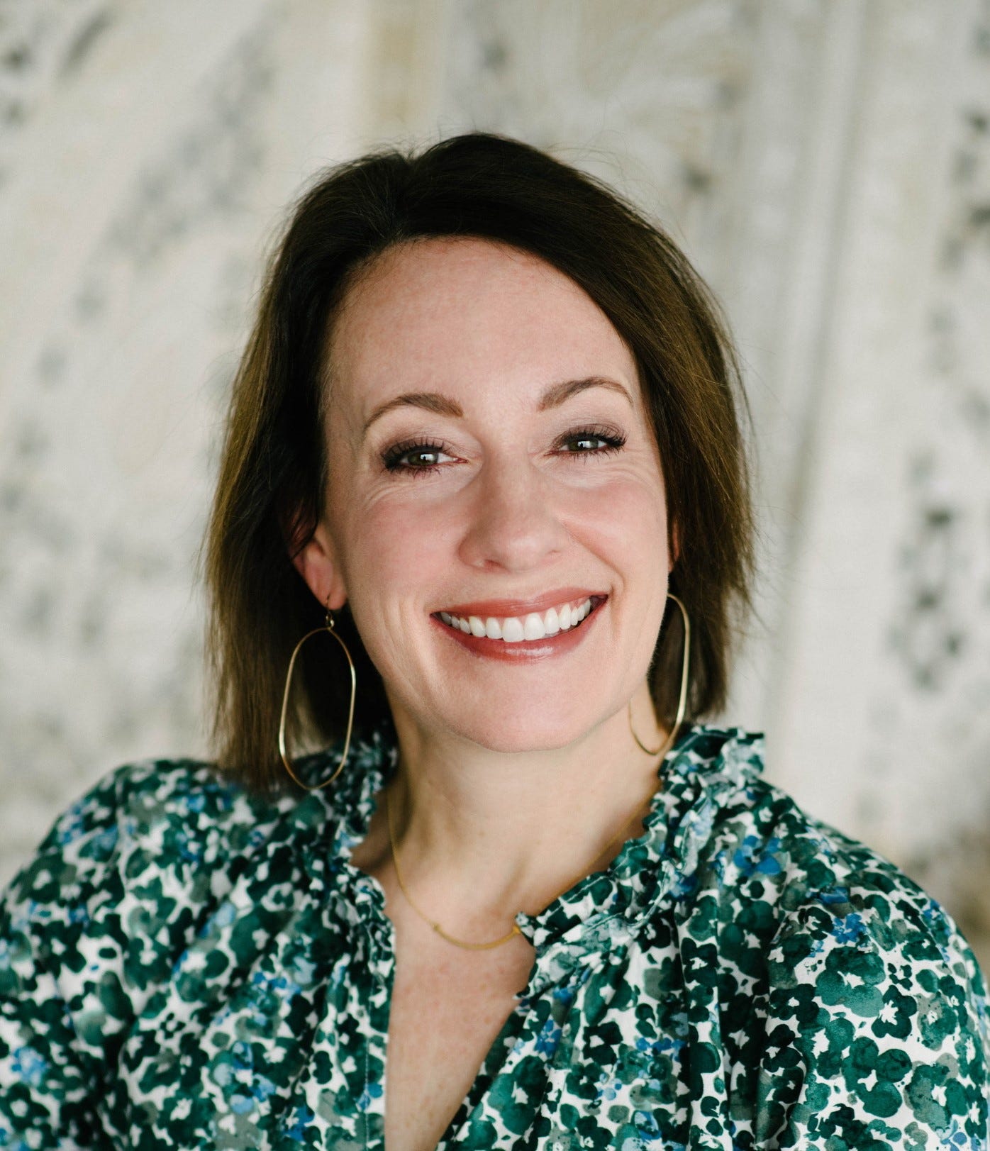 A headshot of a white womean with brown hair, a green flowered blouse, and big gold oval-shaped earrings