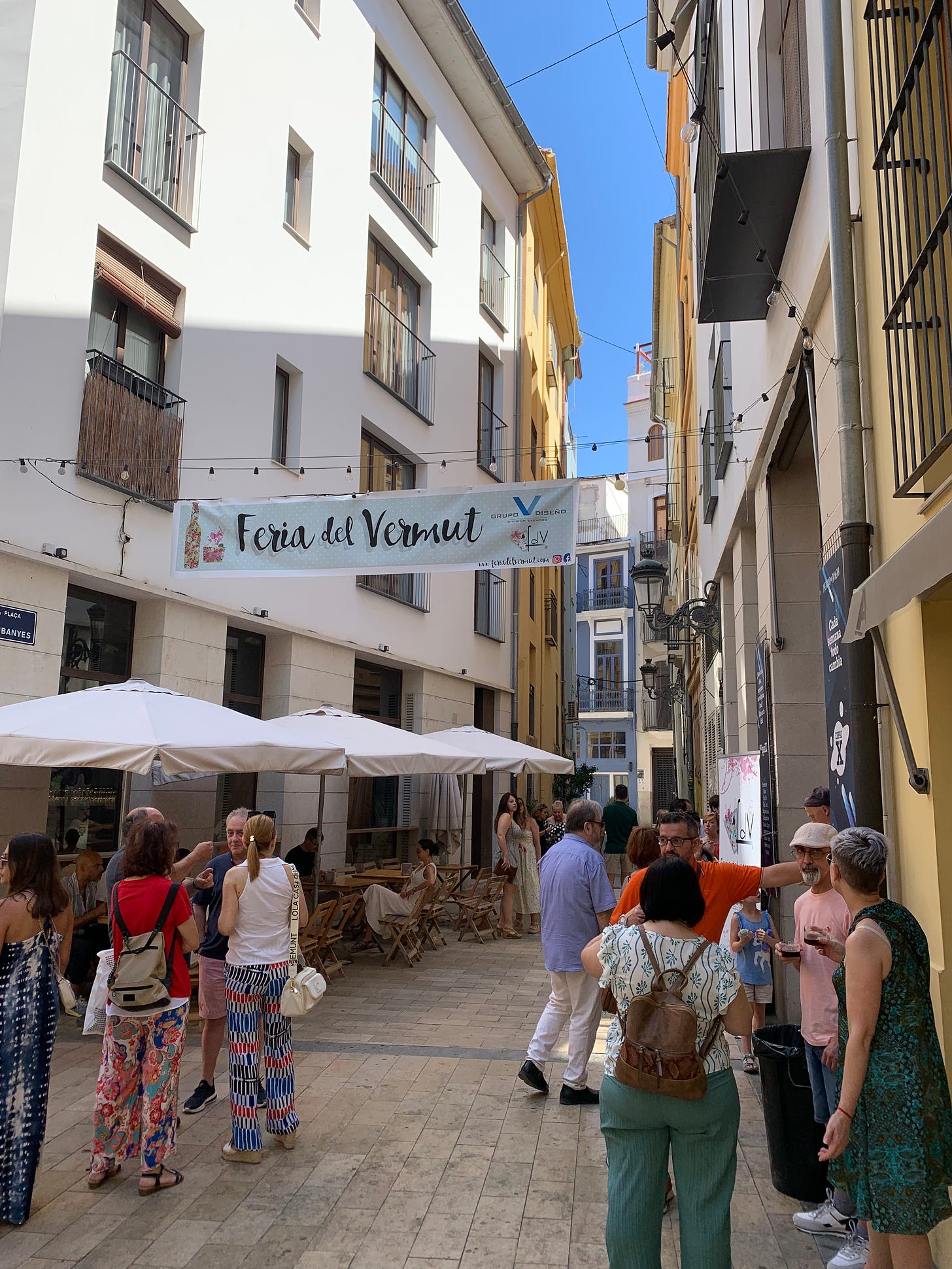 A narrow pedestrian street with a "Feria del Vermut" banner stretched across it and people enjoying glasses of vermut 