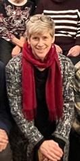 Smiling White woman with grey/red/blond hair, wearing deep red scarf and black/white crazy print blazer, kneeling for family photo with hands crossed on left knee.