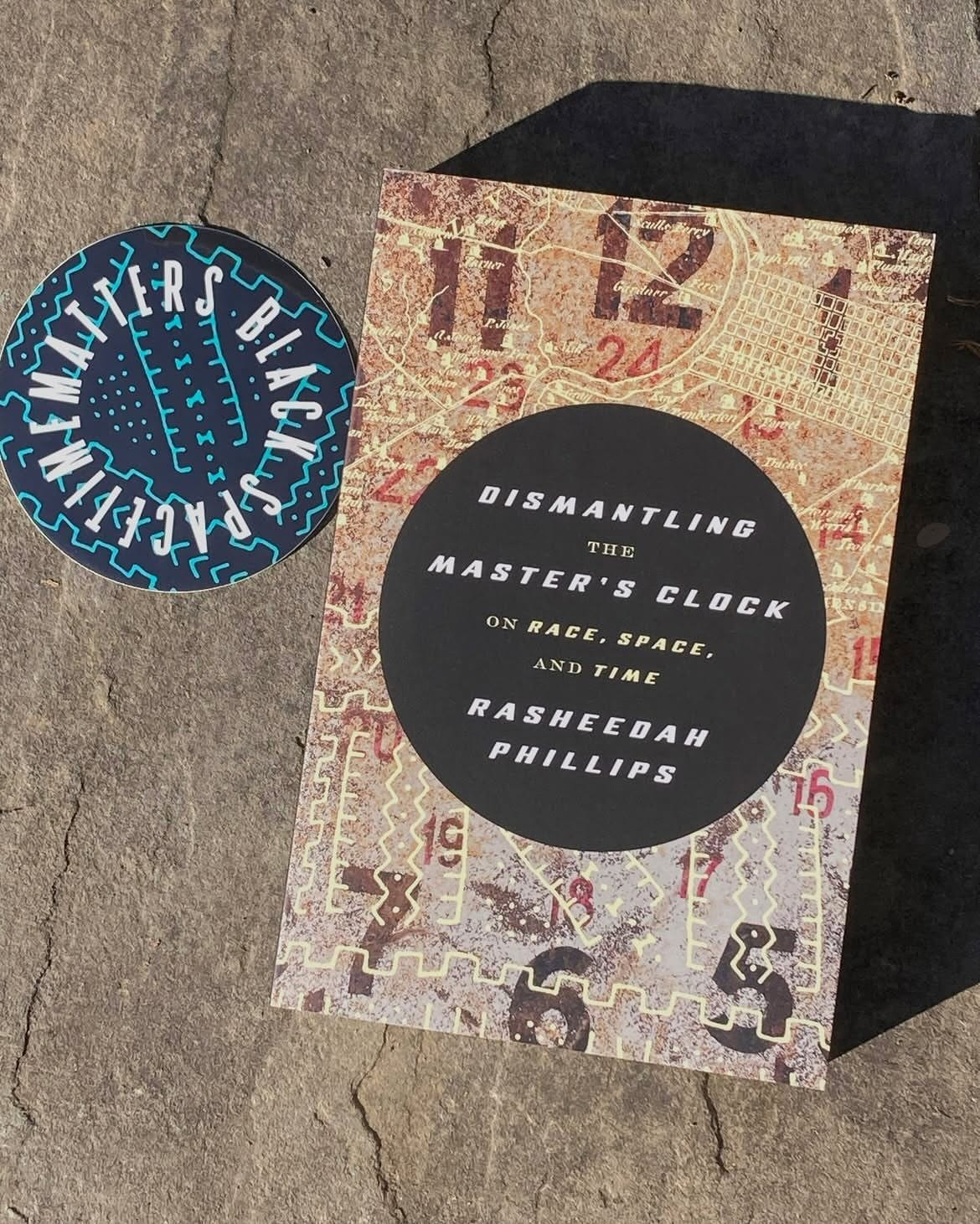 Rasheedah's Phillips new book, Dismantling the Master's Clock, sits on pavement in the sunlight next to a round coaster that says Black Spacetime Matters