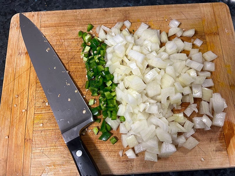 A knife lies on a chopping board next to onion and jalapeno.