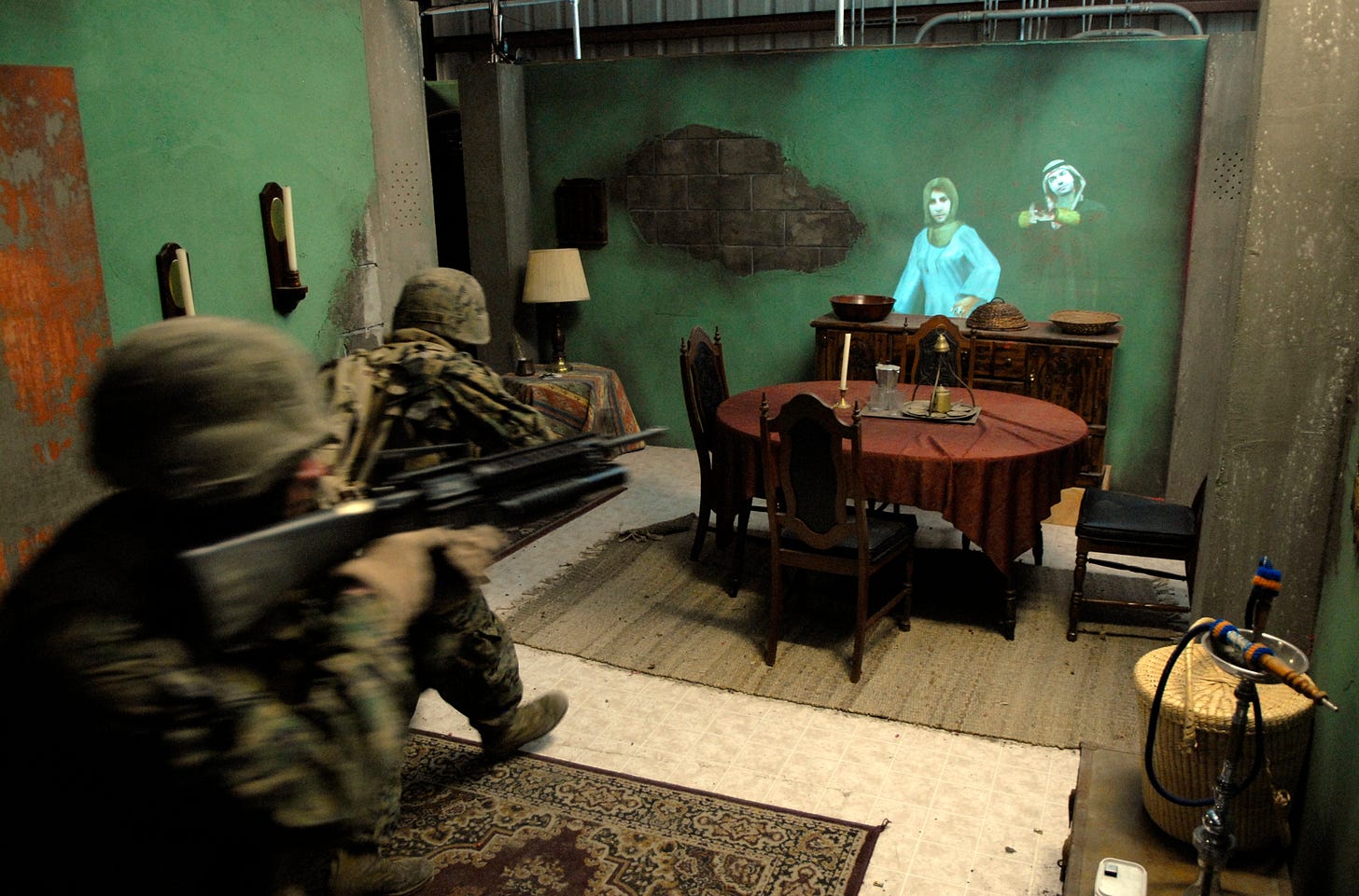 Marines simulate an operation in a staged room, with a dining table, and avatars projected on a wall.