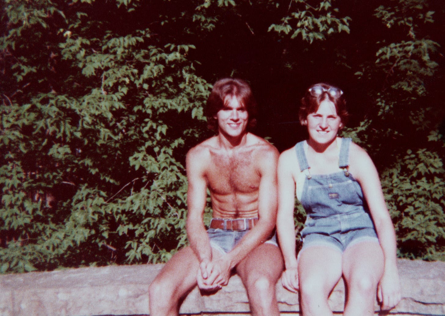 A picture of me—a bare-chested man wearing only cut-off jeans with a wide-ish belt—on the left, and a woman wearing blue jean bibs-shorts. We're sitting on a decorative stone wall in a park near a lake. She has her sunglasses on top of her head. Both of us have hair just below our ears, both parted pretty much in the middle. We are in front of a thicket of bushes and trees.