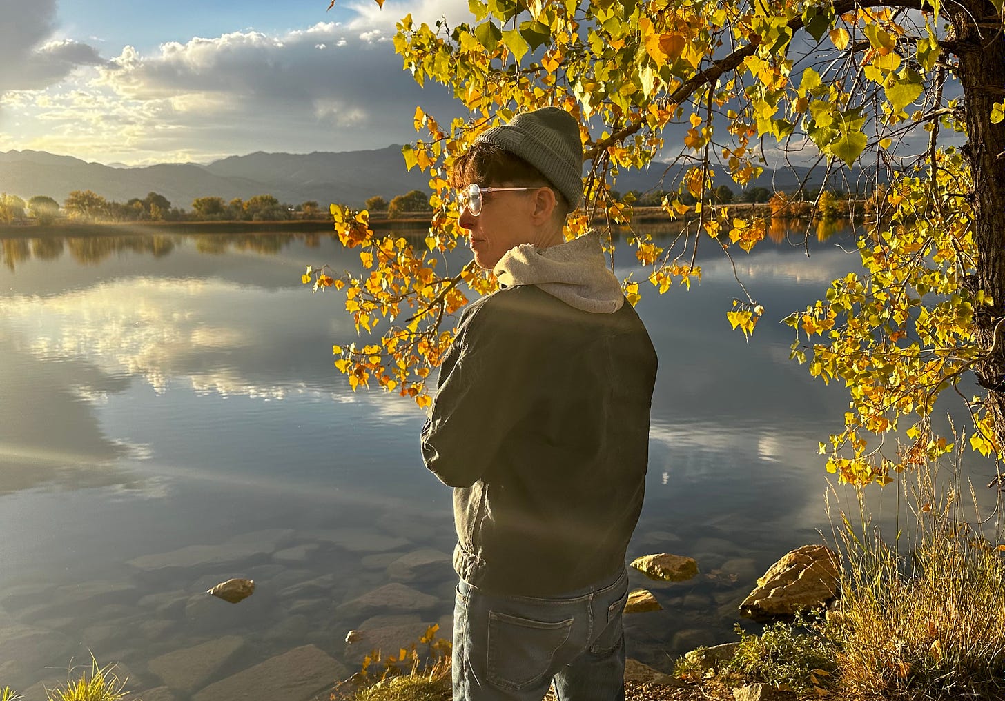 Andrea Gibson standing in front of a lake with mountains, blue skies and scattered clouds in the background and a bright yellow leafed tree next to them. Andrea is wearing a green beanie, sunglasses a green jacket and jeans and pensively looking off into the distance.  