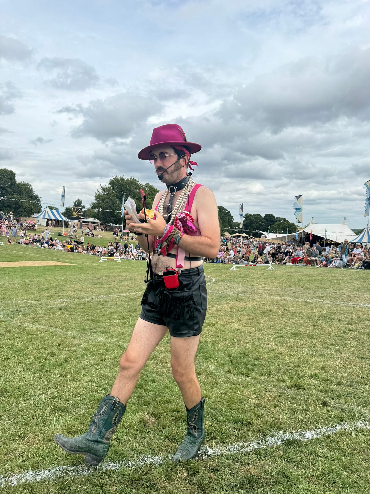 On a playing field with large audience.  A man is wearing some kind of shorts and braces get up.  He is not wearing a shirt.  He has a hat on and is playing the seedy betting man at a horse race.