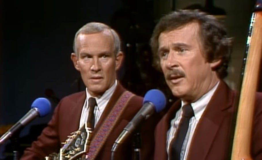 Video screenshot of Tom and Dick Smothers; Tom looks at Dick in pretended worry as Dick explains the song. 