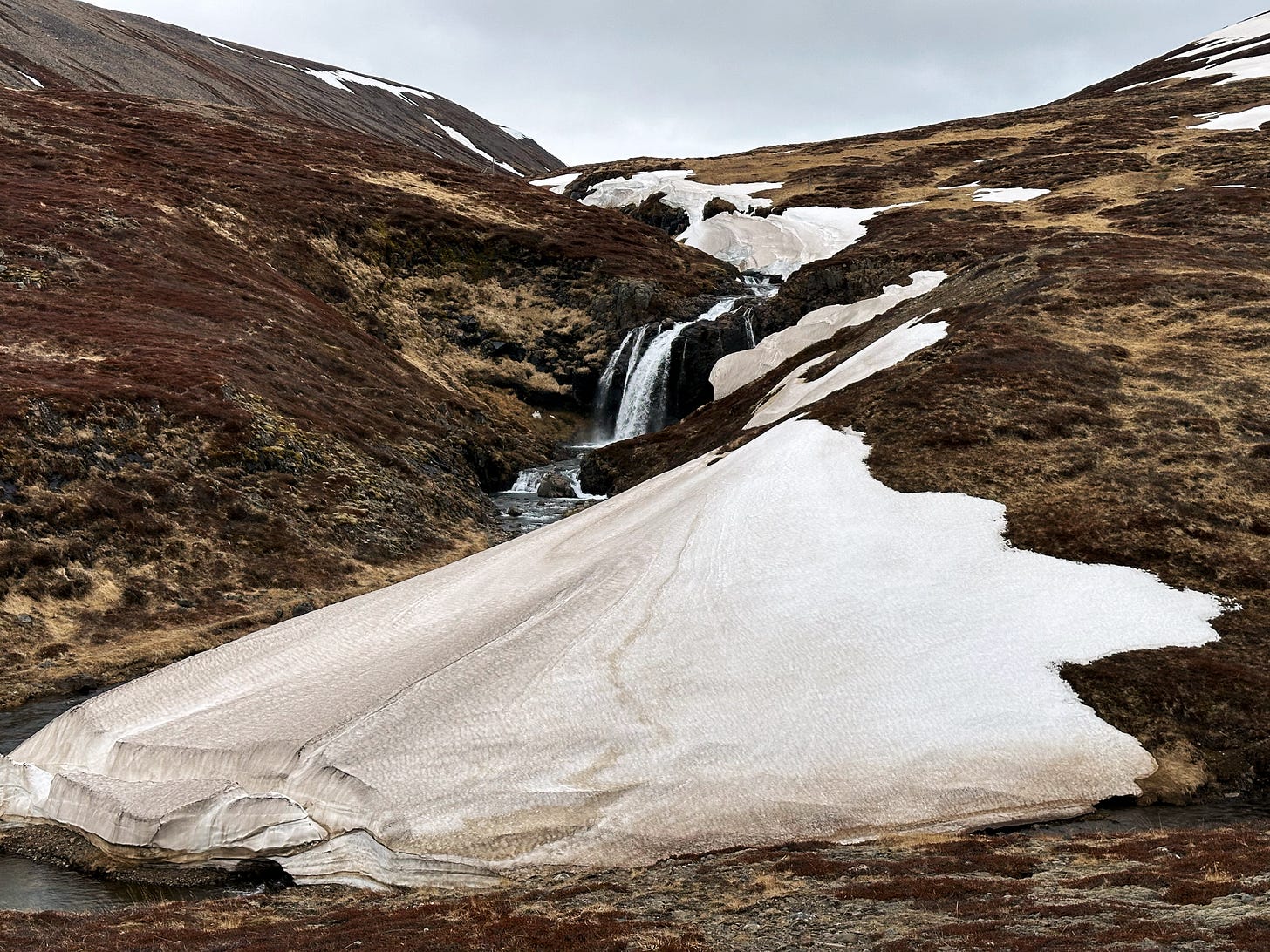 A waterfall cascading through a small valley with winter-brown moss on each side along with white snowdrifts on the right.