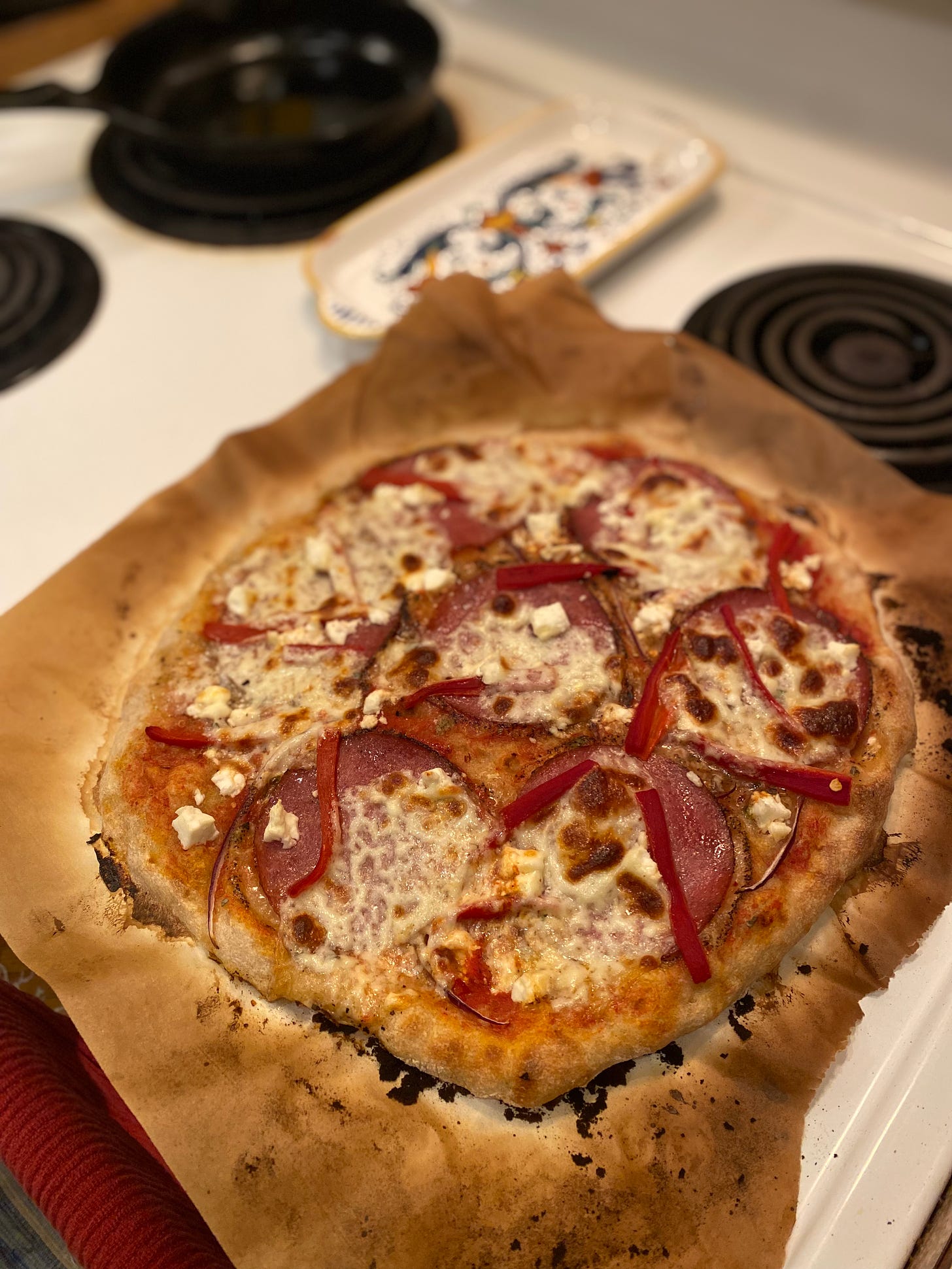 The pizza described above: slices of salami and roasted red pepper and browned cheese over a tomato-sauced crust, set on a piece of parchment burned at the edges from being in the oven. 