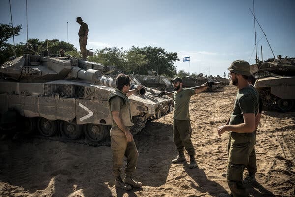 Three men in olive green clothing stand near armored vehicle. The ground is sandy and other armored vehicles are in the background, one flying an Israeli flag.