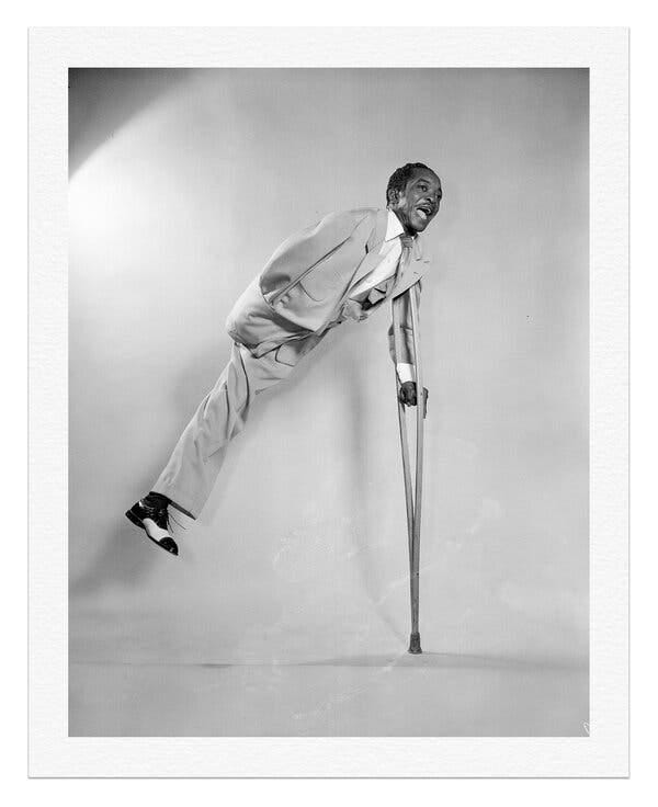 A black and white photo of Henry Heard leaping off a floor, using his crutch with his left arm for balance. He is wearing a light-colored suit and an exuberant expression.