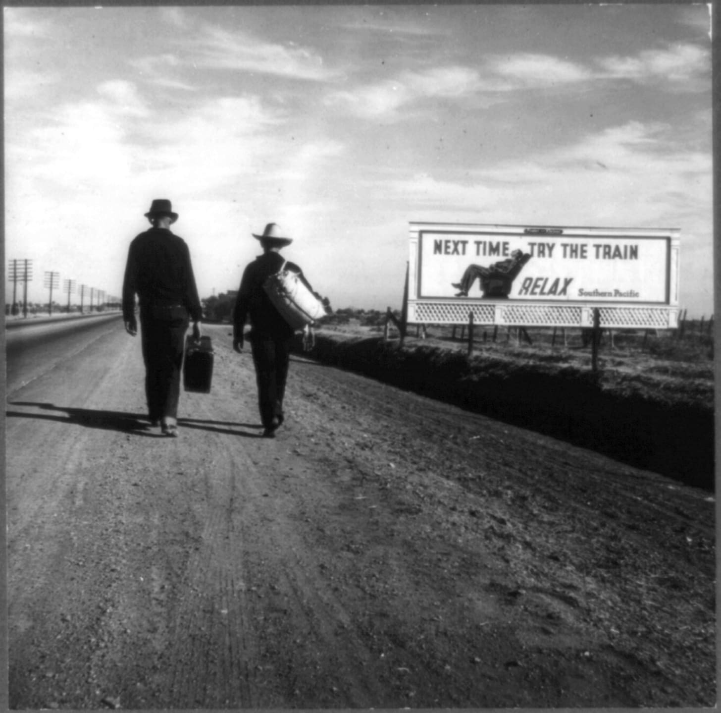 Photo shows two people walking (hitchhiking?) along road near a billboard that says "Next time, try the train. Relax." 