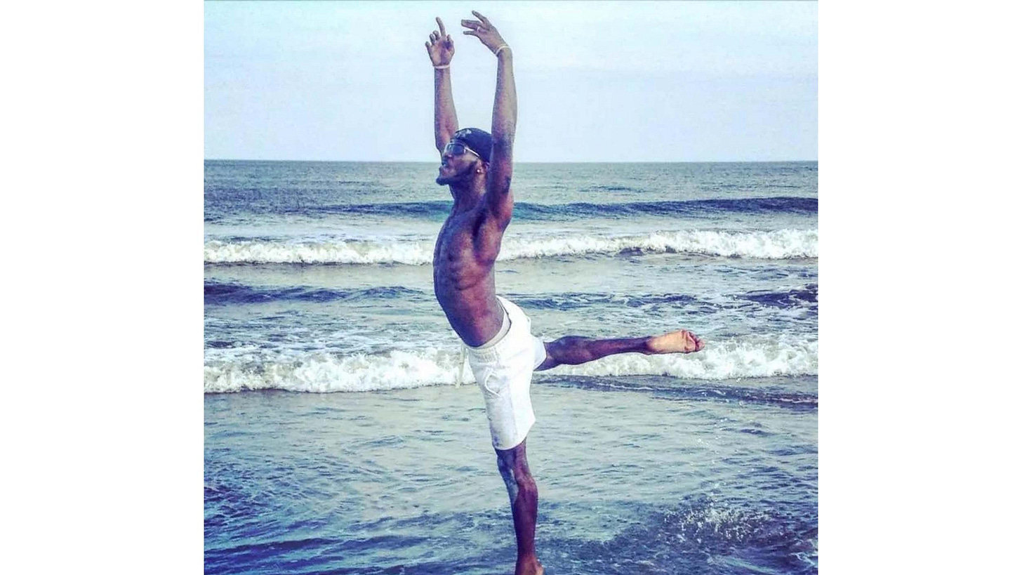 An AfricanAmerican man dancing ballet in shallow water at the beach. He is wearing white shorts and a black hat with...