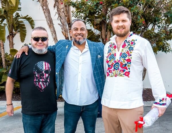 Three smiling men pose together for a photograph outside in front of trees.