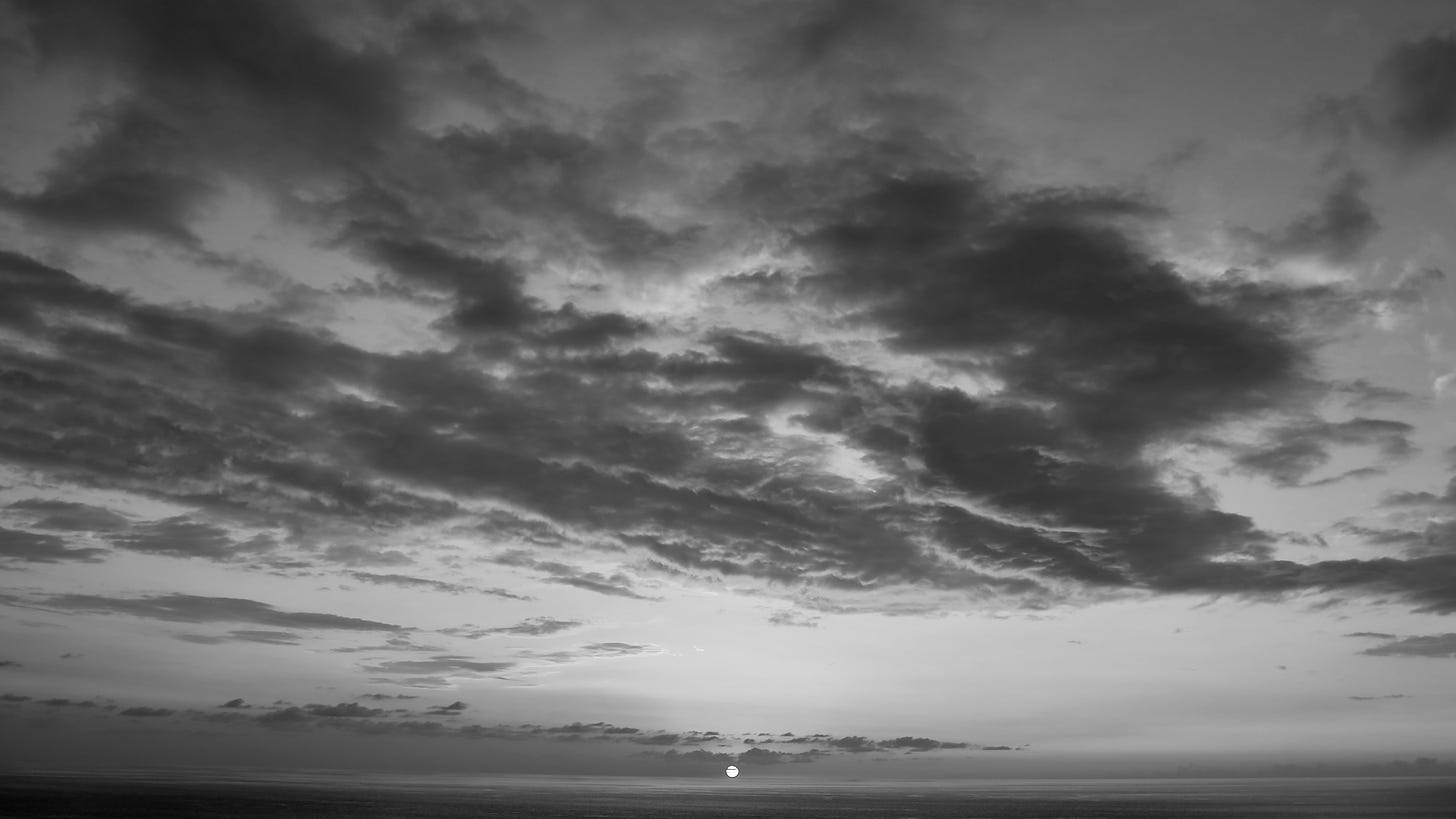 A black-and-white photo from an elevated vantage point of the sun setting over the ocean and illuminating the undersides of clouds above.