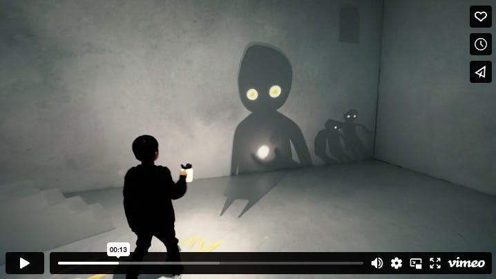 A still from a video showing the art installation 'Chasing Stars in Shadow'. A boy in the foreground shines a lamp, while a shdowy figure on the opposite wall of a grey room holds a ball of light