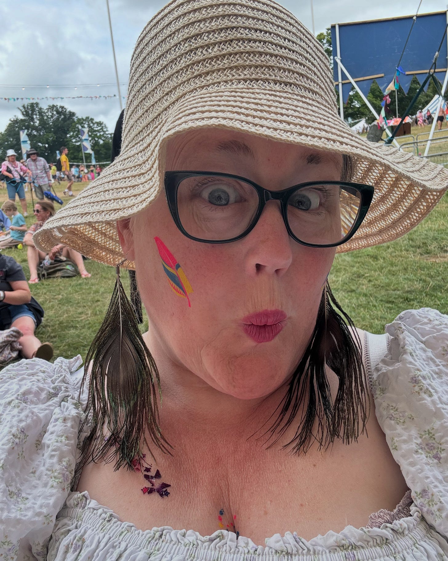 I have a straw hat on and white dress.  I have green glasses.  Glow in the dark sticker tattoos of feathers on my cheeks and chest.  I am making an  'oo' sound with my mouth.