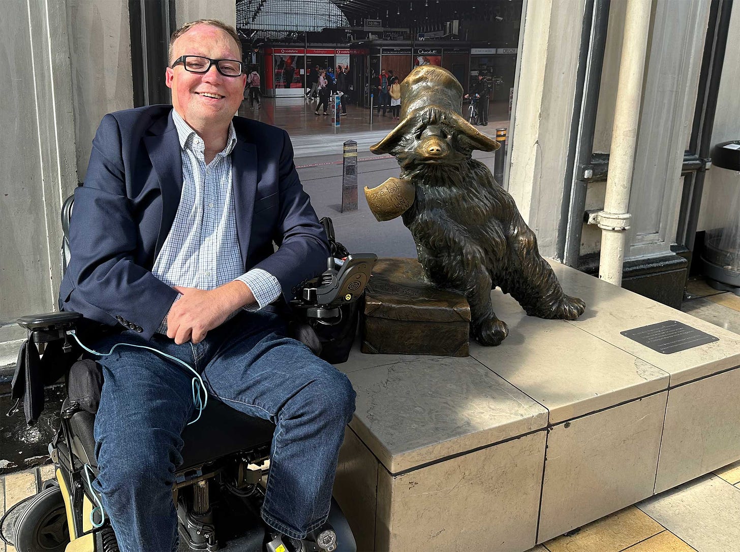 John seated in his wheelchair next to a statue of the Paddington Bear.