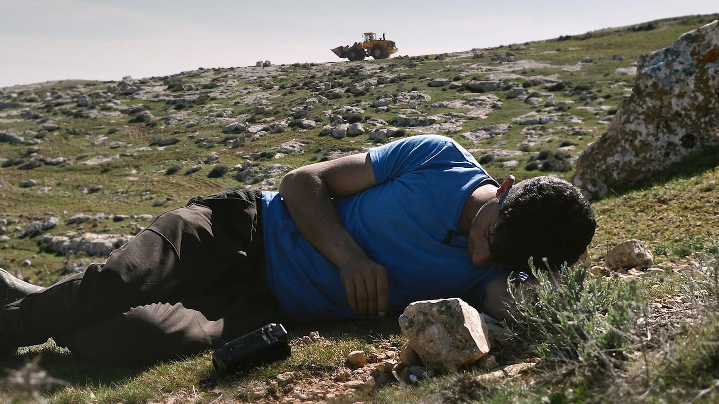 Movie still from No Other Land. A man lies down on the ground, a tractor in the distance on the hill.