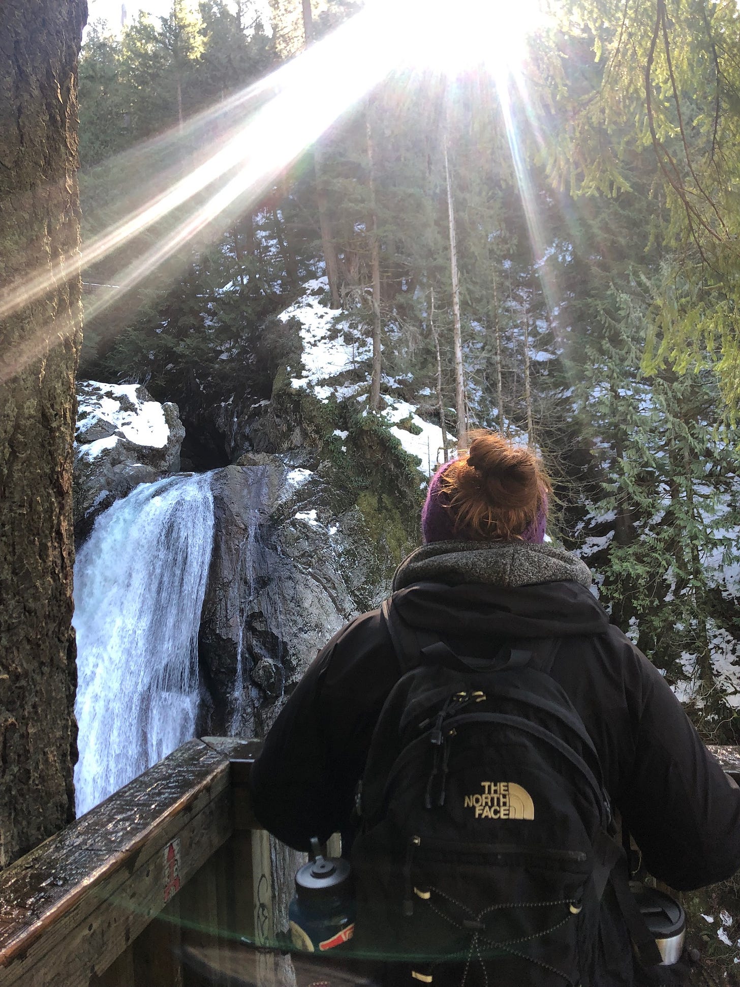 Kate stands with her back to the camera on a snowy trail facing a frozen waterfall while the sun shines through the trees